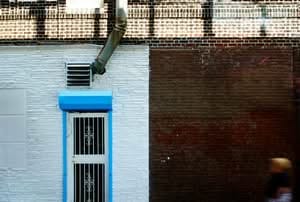 A building exterior vent covers wall divided vertically between white textured stone and dark red brick, featuring a blue-framed security door, an industrial exhaust fan, and a metal downspout.