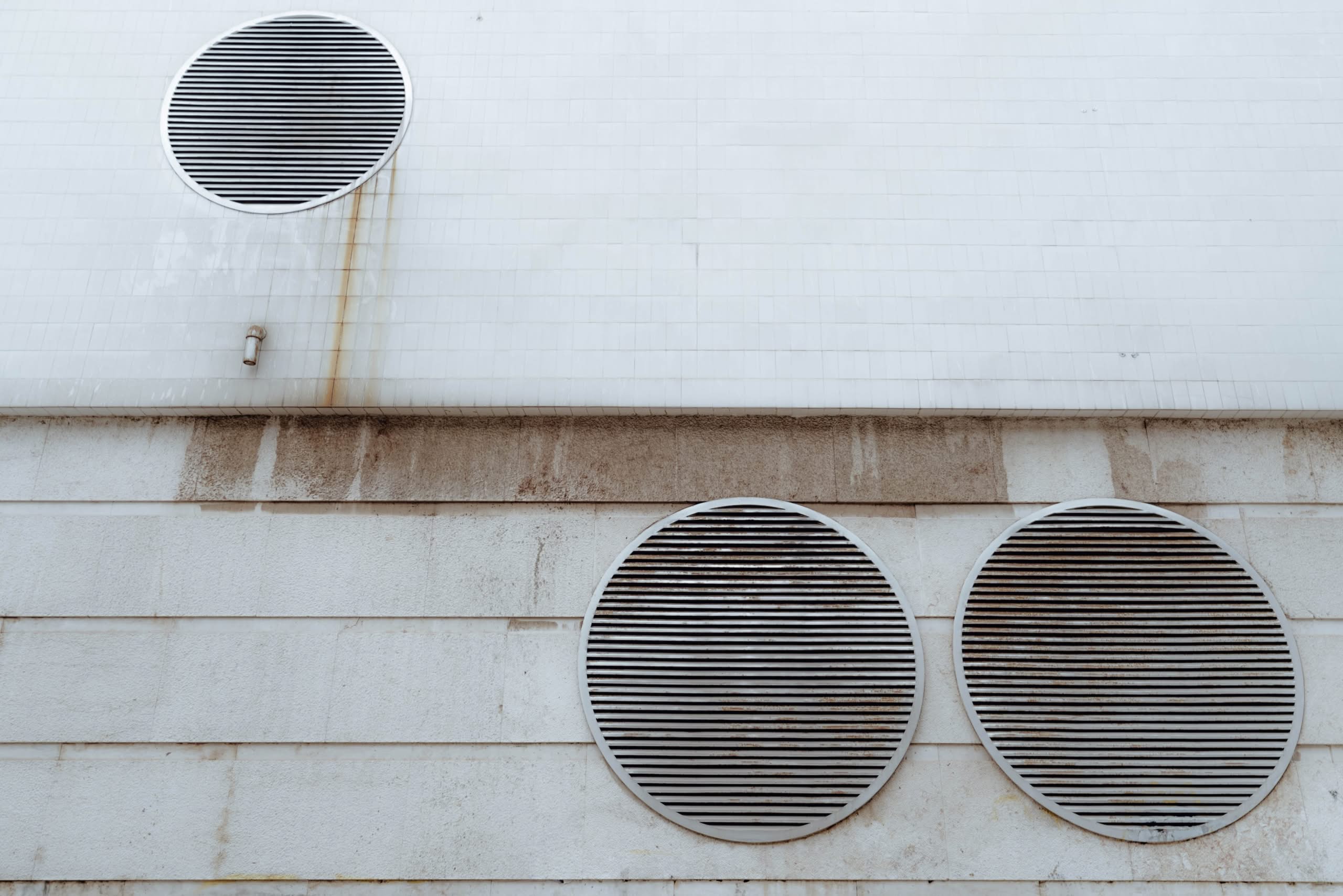 Multi-Story Dryer Vent Exhaust Guide for Safety & Cleaning 1 A white tiled and concrete exterior wall featuring three large circular metal ventilation grilles with horizontal slats, showing minor weathering and rust streaks.
