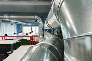 A perspective view of large, shiny silver galvanized steel ventilation long duct runs and spiral pipes hanging from the ceiling of an industrial or modern office space.