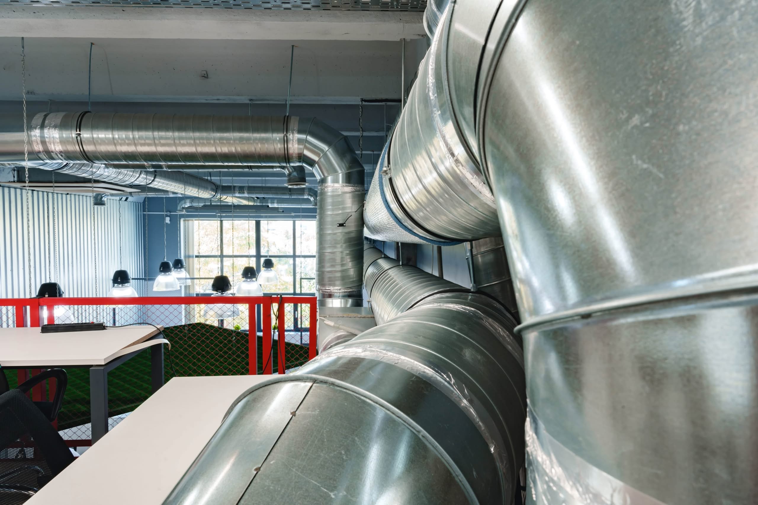 Long Duct Runs in Lady Lake: Risks & Cleaning Guide 1 A perspective view of large, shiny silver galvanized steel ventilation ducts and spiral pipes hanging from the ceiling of an industrial or modern office space.