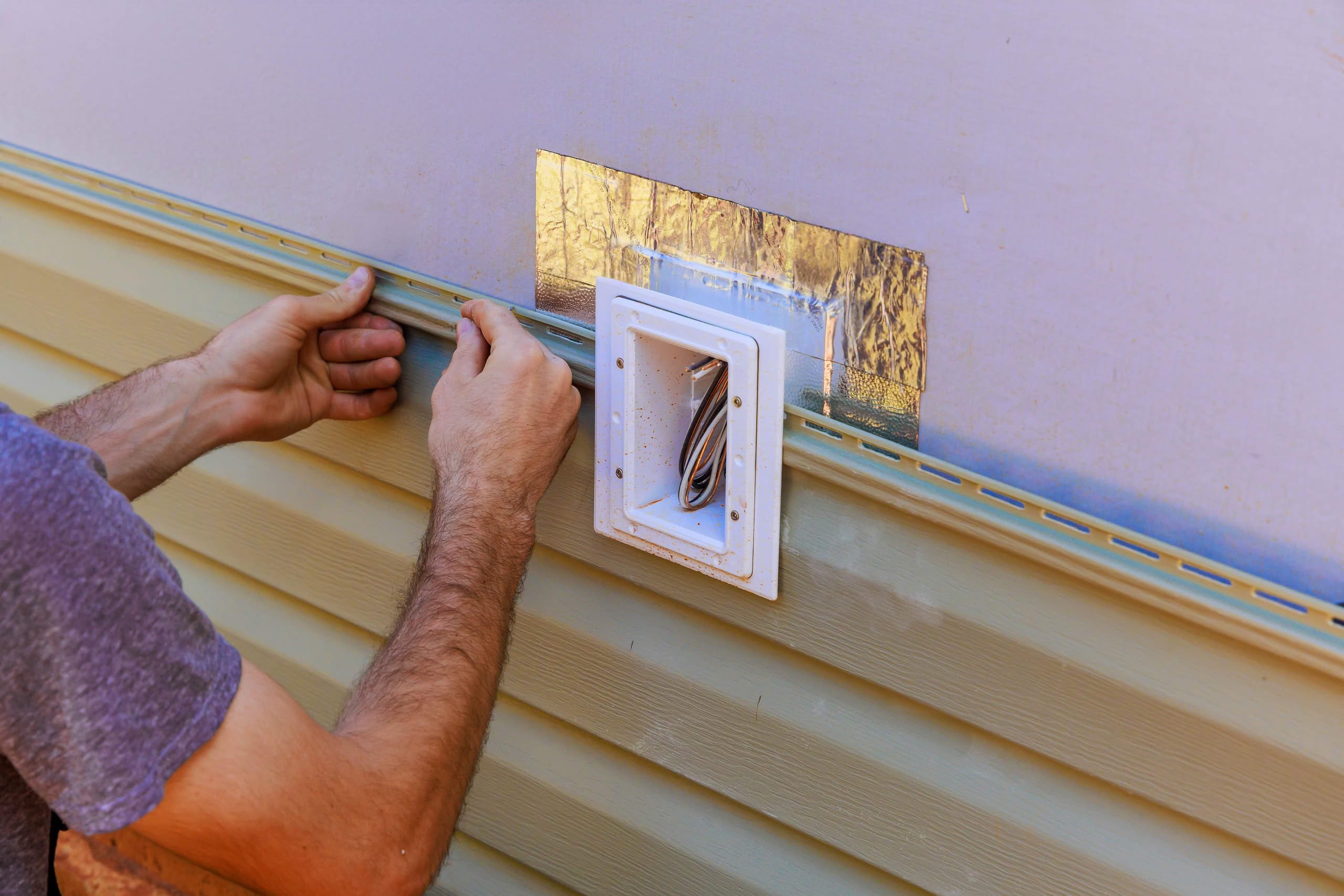 Recessed Dryer Vent Box Installation Ocala: The Ultimate Best Success 1 A close-up of a worker's hands precisely fitting a tan vinyl siding panel around an electrical outlet box on the exterior wall of a house during a renovation project.