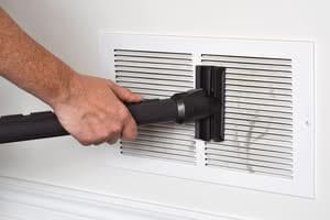 A person's hand using a black vacuum cleaner attachment to remove trapped moisture in vents and lint from a white slatted return air vent on a wall.