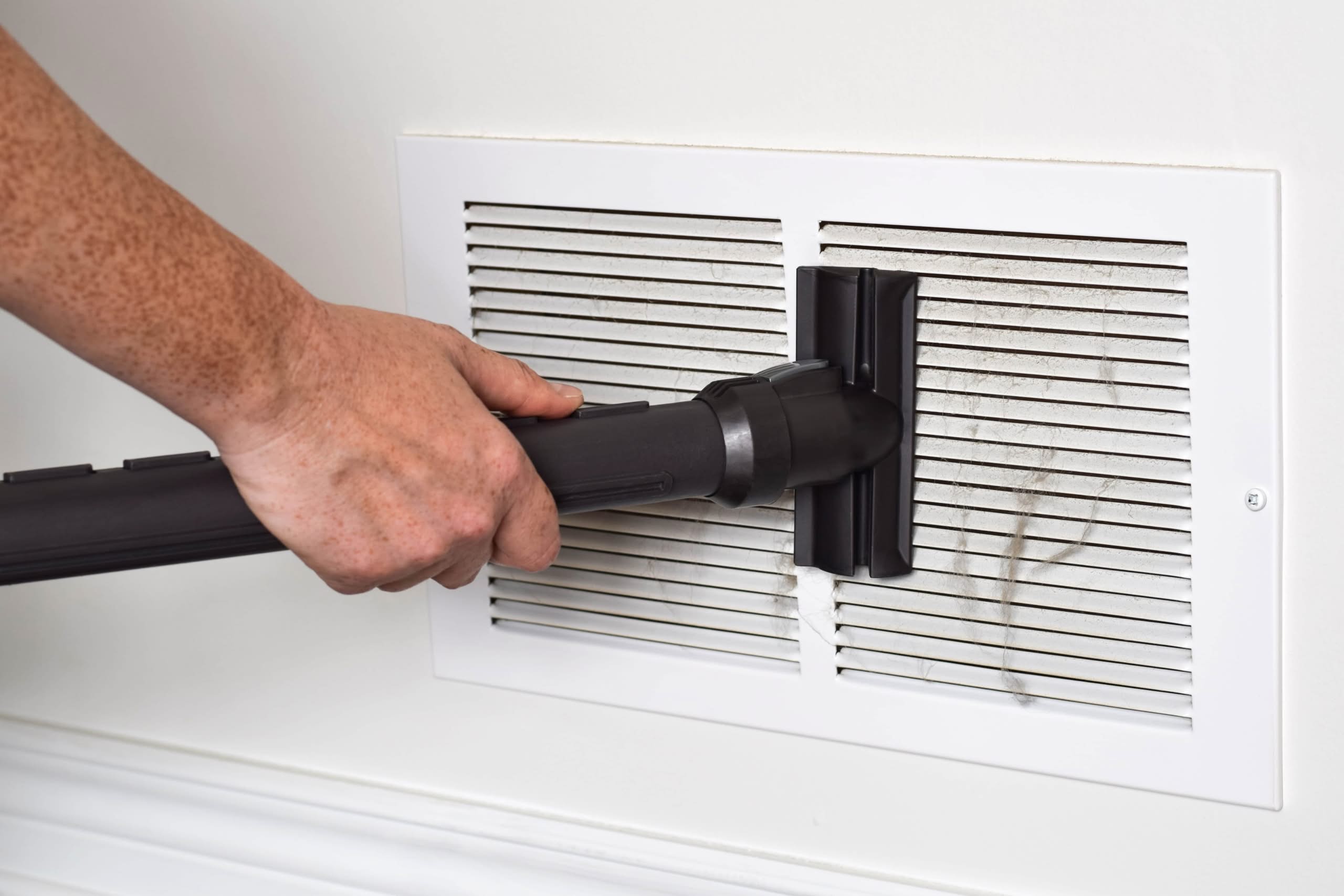 Trapped Moisture in Vents in Clermont Mold & IAQ Guide 1 A person's hand using a black vacuum cleaner attachment to remove thick gray dust and lint from a white slatted return air vent on a wall.