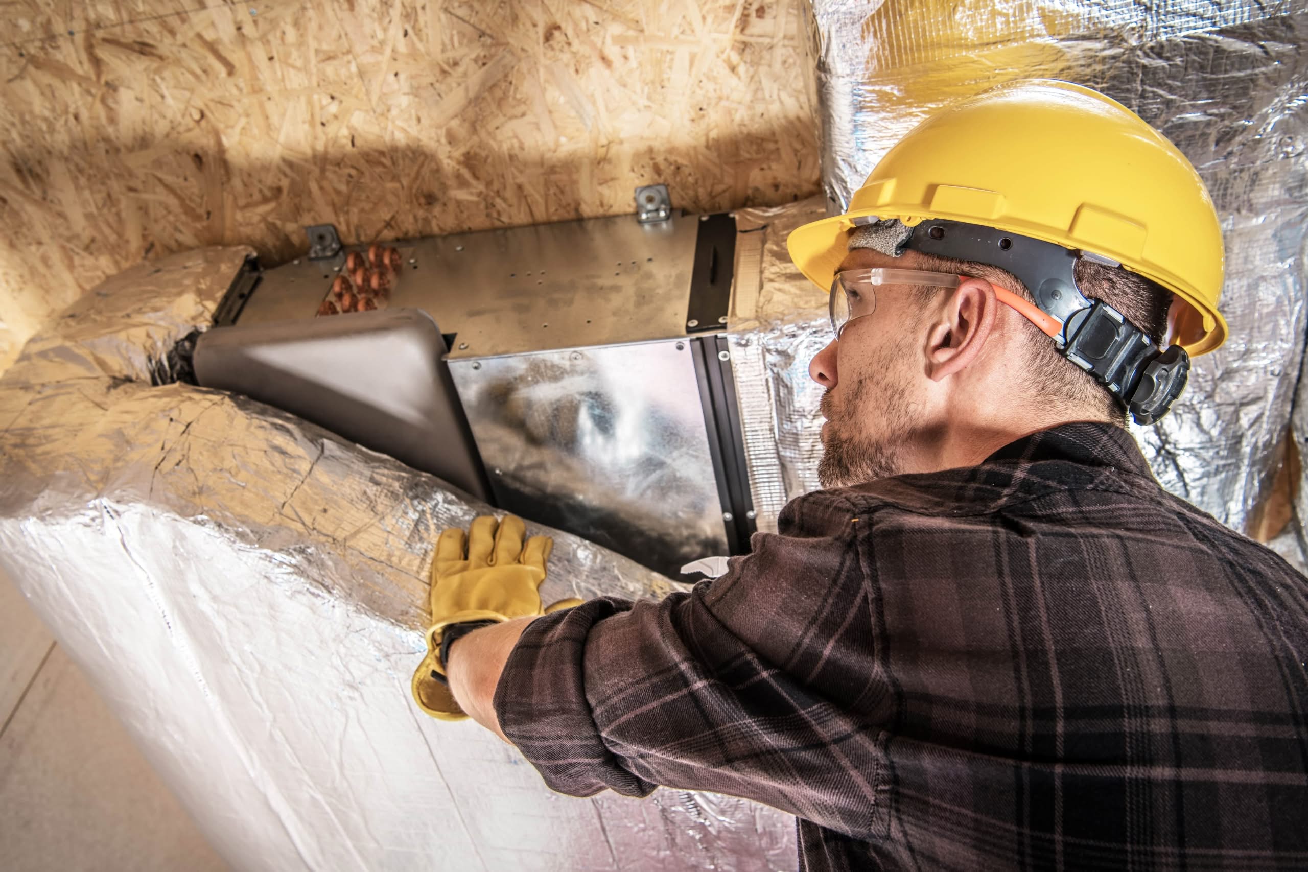 Stop the Humidity: Upgrading Your Exterior Vent Terminations in Citrus County 1 An HVAC worker in a yellow hard hat and safety glasses applying silver foil insulation wrap to metal air ducts in a residential attic.