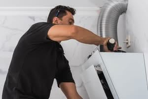 A certified dryer vent cleaning in a black shirt using a power drill and a spirit level to install a white modern kitchen cabinet against a marble-tiled wall near a silver ventilation duct.