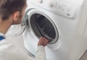 Over-the-shoulder view of a repair technician inspecting the rubber door seal and drum of a white front-load washing machine for mold or wear and dryer vent safety inspections.