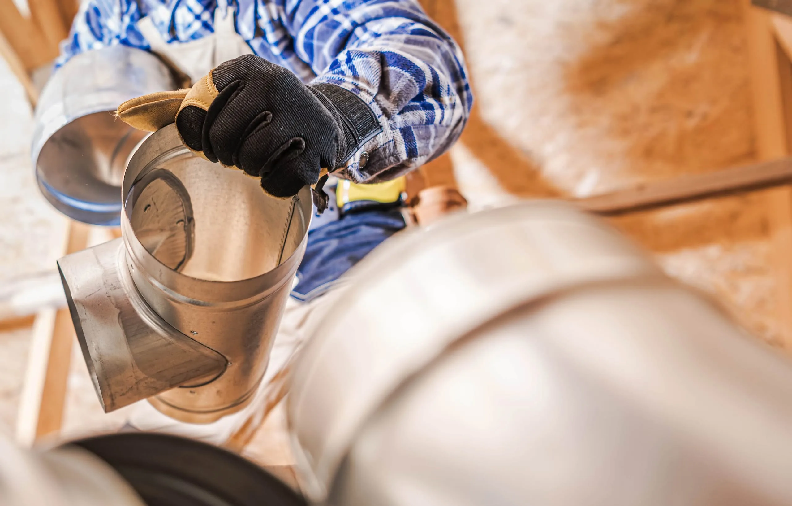 Dryer Vent Rerouting for Better Airflow Leesburg: The Ultimate Proven Solution 1 Close-up of a worker in a plaid shirt and work gloves assembling galvanized steel round ductwork and elbow joints in a residential attic space.