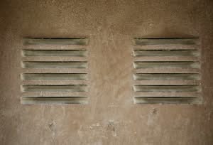A close-up view of two older dryer vents built into a textured, dusty beige exterior stucco wall in Las Vegas, Nevada, showing accumulated dirt around the slats.